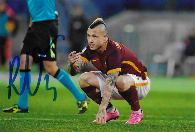 Fotografia autografata Radja Nainggolan accovacciato in campo maglia Roma bordeaux Nike dettagli arancioni tatuaggi braccia gambe mohawk bicolore scarpini rosa fluo arbitro sullo sfondo – autografo originale pennarello blu lato sinistro – 15x10,5 cm – condizioni Excellent