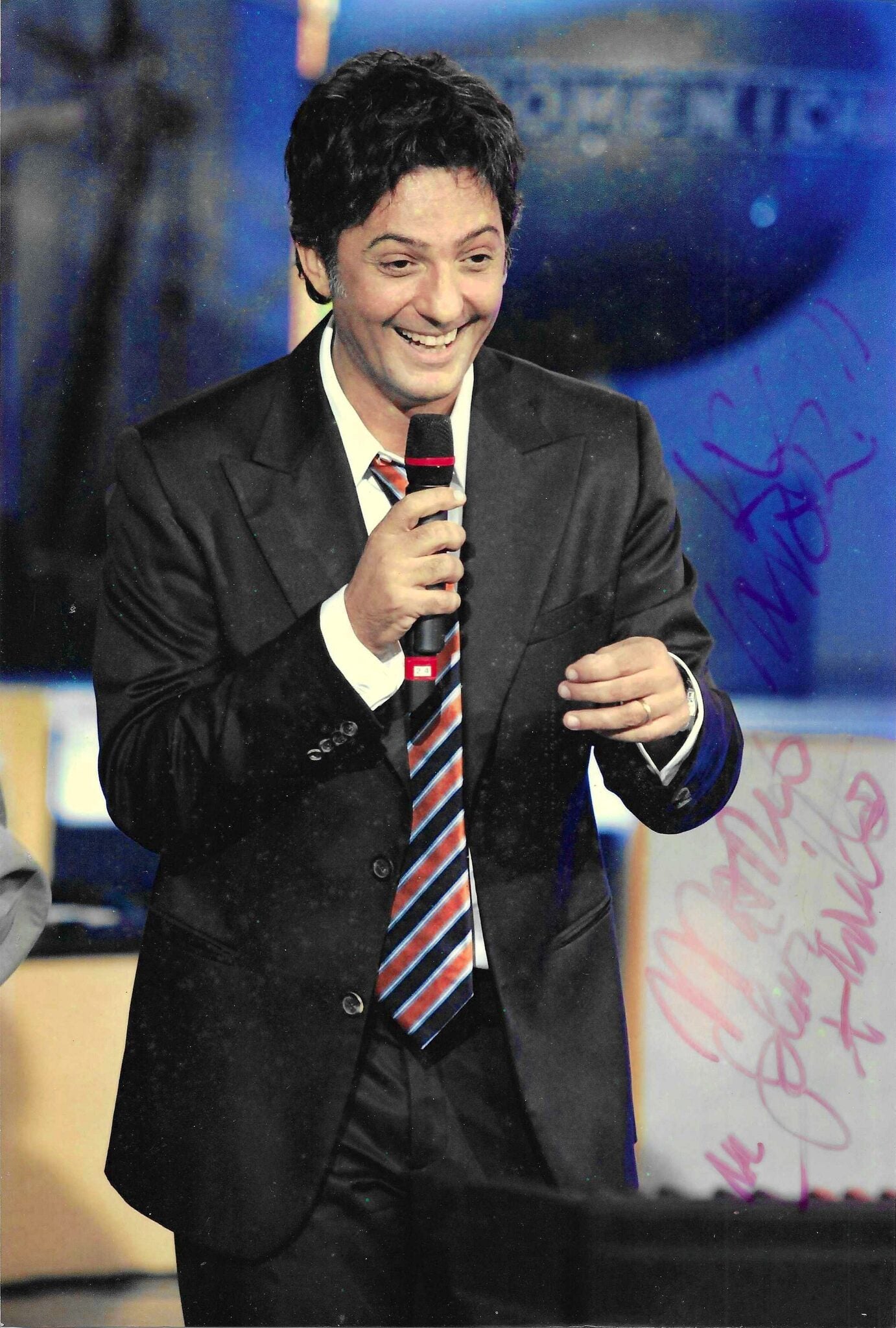 Fotografia autografata con dedica Rosario Fiorello sorridente microfono in mano abito nero cravatta righe rosse blu palco studio televisivo Domenica In RAI – dedica autografa originale pennarello fucsia lato destro – 29,5x20,5 cm – condizioni Excellent