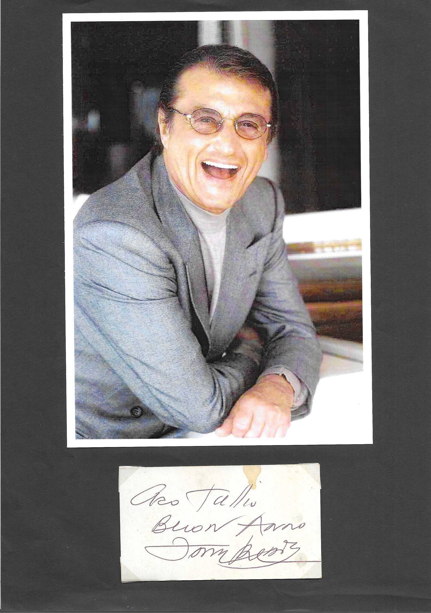 Ensemble autografato Tony Renis fotografia colore sorridente al pianoforte abito grigio occhiali tondi su passepartout grigio con bigliettino autografato dedica Caro Tallu Buon Anno Tony Renis – 9,7x5,5 cm biglietto – condizioni Excellent