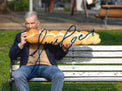 Fotografia autografata Gene Gnocchi comico su panchina mentre addenta una treccia di pane – gag surreale foto Armando Rotoletti – autografo originale pennarello blu