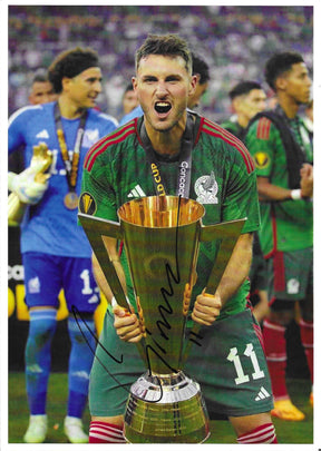 Fotografia autografata Santiago Giménez solleva trofeo CONCACAF Gold Cup urlo di gioia maglia Nazionale Messico verde Adidas numero 11 Ochoa sullo sfondo medaglia oro – autografo originale pennarello nero al centro – 30x21 cm – condizioni Excellent