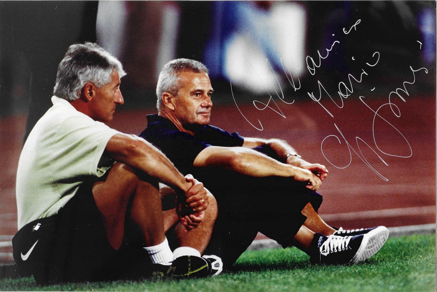 Fotografia autografata con dedica Luigi Simoni allenatore seduto bordo campo con collaboratore tecnico polo scura scarpe Adidas pista atletica sullo sfondo – dedica autografo pennarello bianco in alto a destra – 29,6x19,8 cm – condizioni Excellent